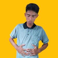 joven asiático hombre participación estómago en dolor vistiendo gris camiseta. aislado amarillo antecedentes. foto