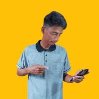 Excited Asian man wearing gray t-shirt pointing to copy space next to holding smart phone, isolated on yellow background. photo