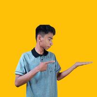 Excited Asian man wearing gray t-shirt pointing at copy space on the side, isolated on yellow background. photo