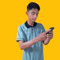 Excited Asian man wearing gray t-shirt pointing to copy space next to holding smart phone, isolated on yellow background. photo