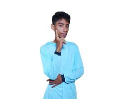 Young thoughtful Asian man holding hand on chin looking pensive, wearing gray t-shirt on blue background. photo