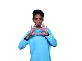 Young Asian man feeling happy and romantic forming heart gesture, wearing blue t-shirt on white background. photo