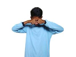 young Asian man crying holding both fists over eyes, wearing blue shirt. isolated white background. photo