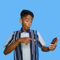 joven asiático hombre sorprendido mirando a inteligente teléfono en izquierda mano participación aislado azul antecedentes foto