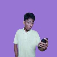 joven asiático hombre sorprendido mirando a inteligente teléfono, vistiendo amarillo camiseta, aislado púrpura antecedentes. foto
