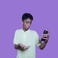 joven asiático hombre sorprendido mirando a inteligente teléfono, vistiendo amarillo camiseta, aislado rosado antecedentes. foto