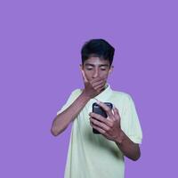 Young asian man surprised looking at smart phone, wearing yellow t-shirt, isolated purple background. photo