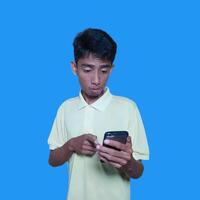 Young asian man surprised looking at smart phone, wearing yellow t-shirt, isolated blue background. photo