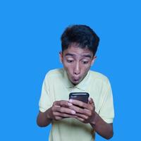 Young Asian man surprised looking at smart phone wearing yellow t-shirt, blue background. photo