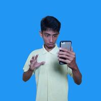 joven asiático hombre sorprendido mirando a inteligente teléfono vistiendo amarillo camiseta, azul antecedentes. foto