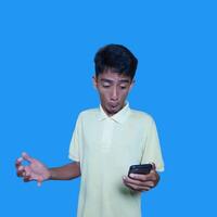 joven asiático hombre sorprendido mirando a inteligente teléfono aislado azul antecedentes foto