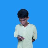 Young asian man surprised looking at smart phone isolated blue background. wearing a yellow t-shirt photo