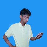 Young asian man surprised looking at smart phone isolated blue background. wearing a yellow t-shirt photo