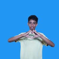 Young Asian man feeling happy and romantic forming heart gesture, wearing yellow t-shirt on blue background. photo