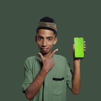 A Young Asian Muslim Man wearing a Skullcap while showing a mobile phone screen isolated on a green background. photo