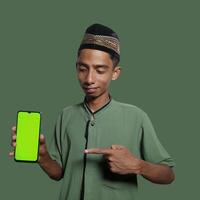 A Young Asian Muslim Man wearing a Skullcap while showing a mobile phone screen isolated on a green background. photo