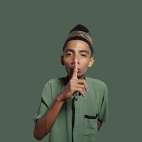 Asian man wearing green muslim shirt on green background, asking for silence with finger on lips. suitable for the Ramadan theme photo