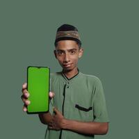 A Young Asian Muslim Man wearing a Skullcap while showing a mobile phone screen isolated on a green background. photo