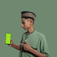 A Young Asian Muslim Man wearing a Skullcap while showing a mobile phone screen isolated on a green background. photo