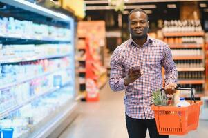 Focused young african american male customer in casual clothes messaging on mobile phone while standing near shelves in grocery shop photo