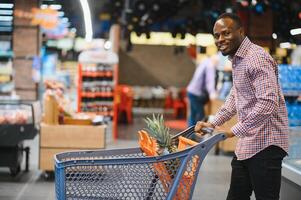 africano americano masculino cliente en casual atuendo con compras carro sonriente y elegir comida desde estantería foto