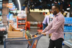 africano americano masculino cliente en casual atuendo con compras carro sonriente y elegir comida desde estantería foto