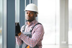 African Workman in overalls installing or adjusting plastic windows in the living room at home photo