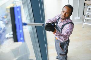 trabajador de limpieza organización cuidadosamente y cuidadosamente frota grande ventana de el oficina espacio. un grave afroamericano en azul mono toallitas el con doble acristalamiento ventana en el oficina foto