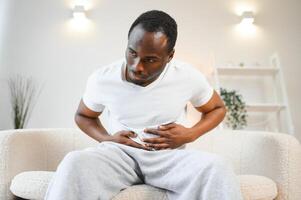 African american guy having stomach ache after eating touching aching stomach suffering from pain sitting on sofa at home. photo