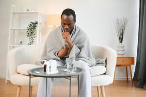 Cold african american young man covered in grey blanket freezing at home, sitting on sofa alone and warming up his hands in living room, copy space photo