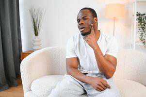 Close-up photo of a young African-American man suffering from a severe toothache at home on the couch. He holds his hand to his cheek, grimacing in pain