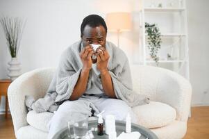 Sick mature black man covered in blanket sneezing runny nose while sitting on couch in living room, with lots of pills on table photo