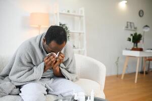 Sick mature black man covered in blanket sneezing runny nose while sitting on couch in living room, with lots of pills on table photo