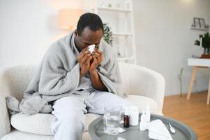 Sick mature black man covered in blanket sneezing runny nose while sitting on couch in living room, with lots of pills on table photo