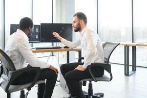 Two diverse colleagues traders talking to each other, sitting in the office in front of multiple computer screens. Stock trading, people, business concept photo