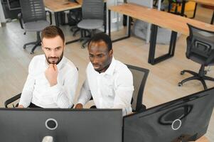 Two diverse colleagues traders talking to each other, sitting in the office in front of multiple computer screens. Stock trading, people, business concept photo