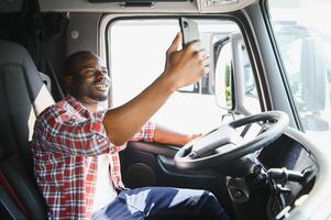 truck driver video call on smartphone in cockpit. Trucker Asian young man beard parked for coffee break after long drive. Worker male driving semi-truck transport logistics express delivery services. photo