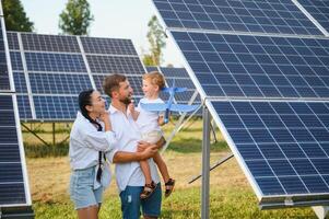 joven familia de Tres es agachado cerca fotovoltaica solar panel, pequeño chico y padres. moderno familia concepto. el concepto de verde energía foto