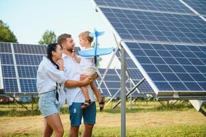 joven familia de Tres es agachado cerca fotovoltaica solar panel, pequeño chico y padres. moderno familia concepto. el concepto de verde energía foto