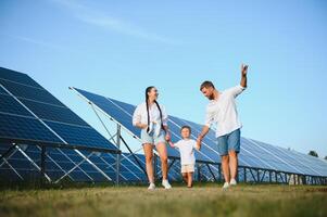 joven familia de Tres es agachado cerca fotovoltaica solar panel, pequeño chico y padres. moderno familia concepto. el concepto de verde energía foto