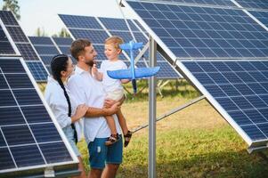 joven familia de Tres es agachado cerca fotovoltaica solar panel, pequeño chico y padres. moderno familia concepto. el concepto de verde energía foto