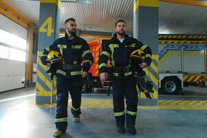 retrato de dos joven bomberos en uniforme en pie dentro el fuego estación foto