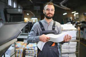 Printing house interior with modern offset print machine and operator in working uniform checking quality and controlling process of print photo