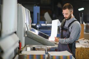 Printing house interior with modern offset print machine and operator in working uniform checking quality and controlling process of print photo