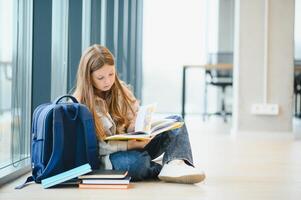 Beautiful schoolgirl smiling while sitting near window and reading book photo