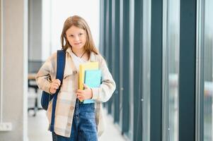 bonito rubia colegio niña participación muchos vistoso notas y libros. inteligente adolescente niña sonriente a cámara, en pie en corredor de internacional colegio foto