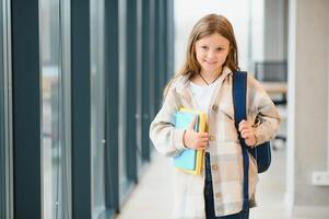 pequeño hermosa colegio niña en pie entre corredor a escuela, participación notas a manos. gracioso y contento niña sonriente a cámara, descansando después lecciones en primario colegio foto