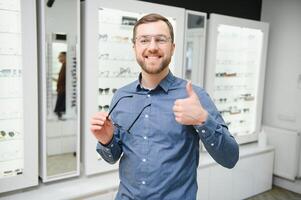 satisfecho cliente. ver de contento joven masculino cliente vistiendo nuevo anteojos, en pie cerca estante y escaparate con anteojos sonriente hombre molesto en gafas foto