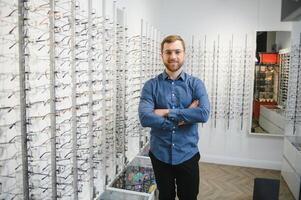 en óptica tienda. retrato de masculino cliente participación y vistiendo diferente gafas, elegir y molesto en nuevo lentes a óptico almacenar. hombre cosecha marco para visión corrección, de cerca. foto