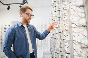 In Optics Shop. Portrait of male client holding and wearing different spectacles, choosing and trying on new glasses at optical store. Man picking frame for vision correction, closeup. photo